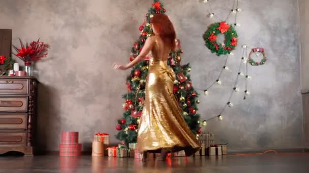 Beautiful woman in golden evening dress spins and dances near Christmas tree — Stock Video