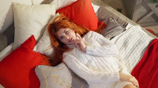 Accogliente donna dai capelli rossi sexy in maglione bianco a maglia si trova sul divano e sorride a casa — Video Stock
