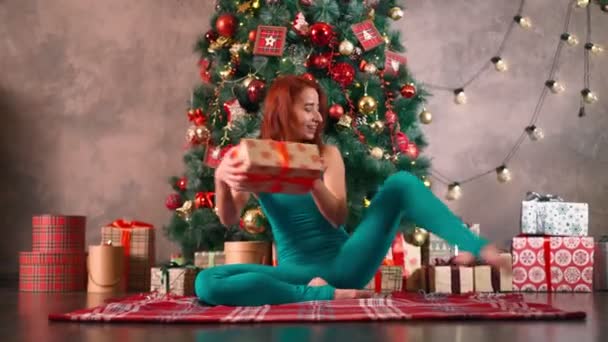 Vrouw zit in splits opent Nieuwjaar cadeau achtergrond kerstboom. — Stockvideo