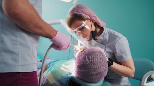 アシスタントや医療機器の助けを借りて保護シールドとフェイスマスクの美しい女性歯科医は 歯科事務所で女性患者で動作します — ストック動画