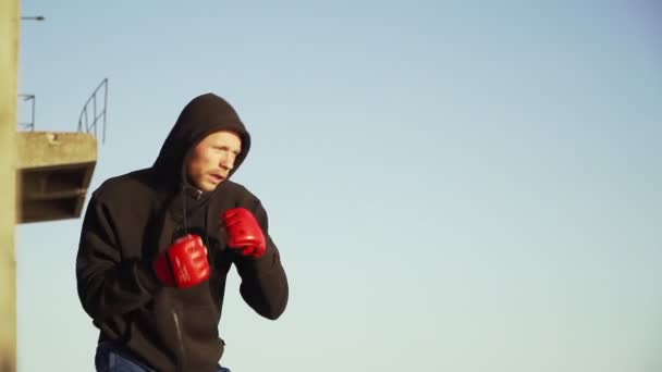 Kaukasischer Boxer in roten Handschuhen im schwarzen Sweatshirt mit Kapuze macht einen Schattenkampf — Stockvideo