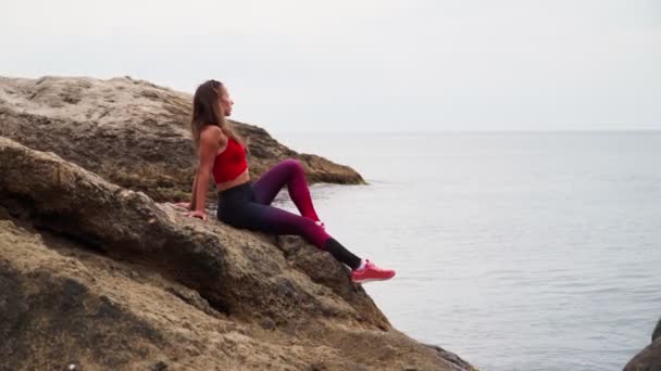 Schöne athletische Mädchen in Sportbekleidung sitzt auf einem Felsen inmitten des Meeres Hintergrund der schönen Landschaft — Stockvideo