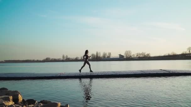 Una ragazza atletica che corre sul molo lungo il lago sullo sfondo di un bellissimo tramonto. — Video Stock