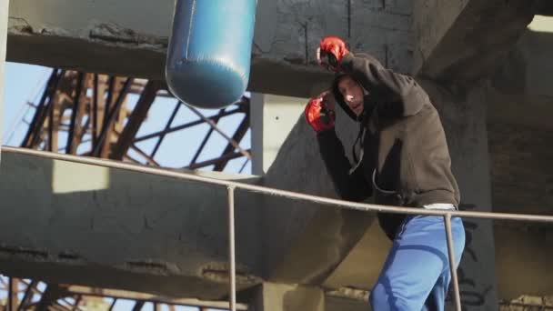 Kaukasischer Boxer schlägt Boxsack im Hintergrund eines verlassenen Gebäudes. Boxen und Kampfkunst — Stockvideo
