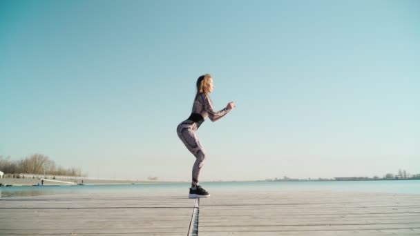 Genç fit esmer güzel kız tozluklu, siyah spor ayakkabılı mavi göle karşı spor egzersizleri yapıyor. — Stok video