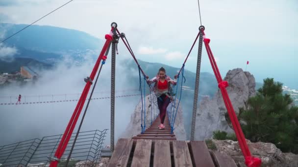 Chica atlética joven es un viajero que cruza el puente de la cuerda por encima del precipicio en las montañas. — Vídeo de stock