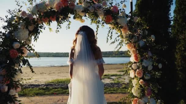 Невеста в свадебном платье стоит под украшенным арочным фоном красивый закат — стоковое видео