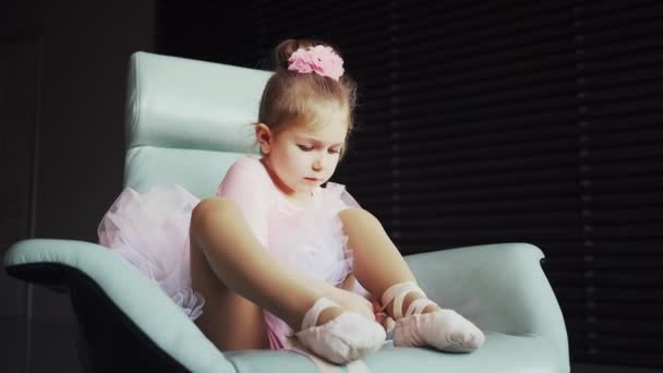 Linda niña en tutú rosa sentada en silla azul y atando zapatos de ballet. De cerca. pequeña bailarina — Vídeo de stock