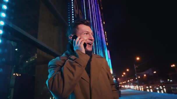 Alışveriş merkezi kış gecesinin dışında cep telefonuyla konuşan ve gülen neşeli adam. — Stok video