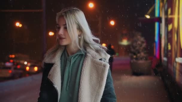 Girl Walks and Smiles Winter Night during Snowfall near Glowing Shopping Center — Stock video