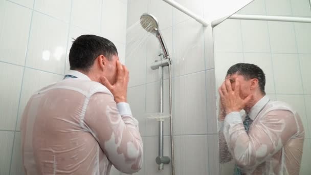 Грустный человек в рубашке и галстуке стоит в ванной комнате под душем и ванны в одежде — стоковое видео