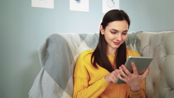 Женщина, сидящая на удобном диване, улыбается и изучает приложение на цифровом планшете — стоковое видео