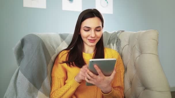 Mujer sentada en el sofá acogedor sonrisas y estudios aplicación en la tableta de ordenador digital — Vídeos de Stock