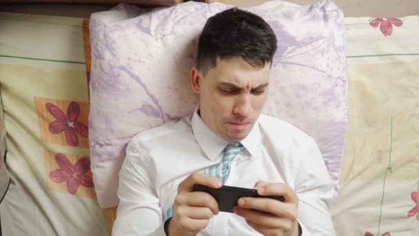 Engraçado homem joga um jogo no telefone e ganha enquanto deitado no sofá em casa. — Vídeo de Stock