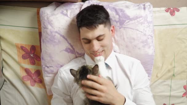Όμορφος νέος με ρούχα γραφείου μετά τη δουλειά, ξαπλωμένος στο κρεβάτι με τη γάτα του. — Αρχείο Βίντεο