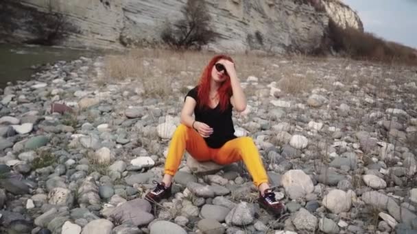 Mujer de moda en gafas de sol sentado descansando en la orilla rocosa del río de montaña — Vídeo de stock