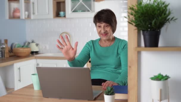Щаслива літня жінка, що спілкується за допомогою відеодзвінка, використовуючи ноутбук вдома на кухні — стокове відео