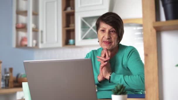 Feliz anciana comunicándose por videollamada usando portátil en casa en la cocina — Vídeos de Stock