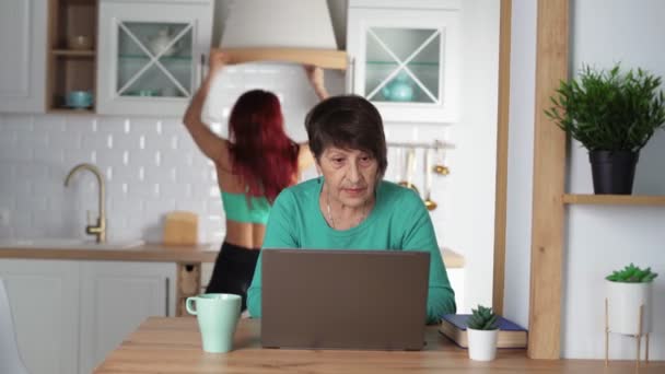 Пожилая женщина удаленно работает на ноутбуке дома, ее дочь танцует Удаленный труд — стоковое видео