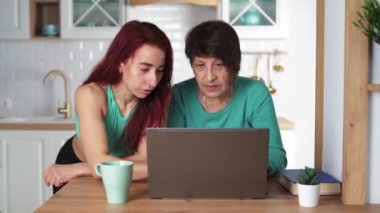 Young girl explains to her elderly mother how to use a laptop at home in kitchen