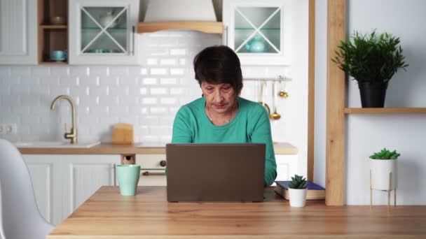 Mujer anciana enfocada remotamente trabajando en la computadora portátil en casa. Personas de edad avanzada — Vídeos de Stock