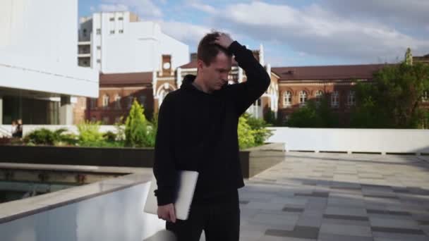 Muž na volné noze stojí s laptopem v rukou a narovnává si vlasy — Stock video