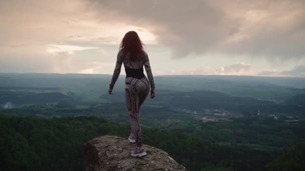 Стройная девушка стоит на вершине горы и поднимает руки, ошеломляя пейзаж — стоковое видео