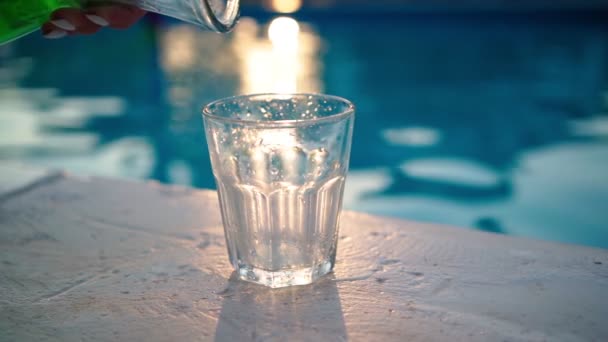 Green refreshing drink is poured from bottle into glass backdrop swimming pool. — Stock Video