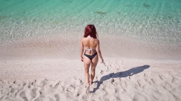 Красивая девушка с рыжими волосами в купальнике выходит в прозрачное бирюзовое море — стоковое видео
