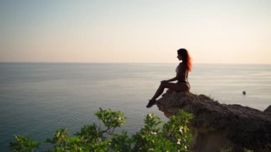 Young Slender Woman in Swimsuit Sits on Rock backdrop Sunset and Seascape