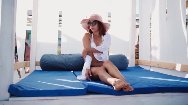 Mujer delgada en sombrero se sienta relajante y disfruta tomando el sol en gazebo en la playa de mar — Vídeos de Stock