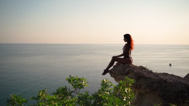 Young Slender Woman in Swimsuit Sits on Rock backdrop Sunset and Seascape — Video