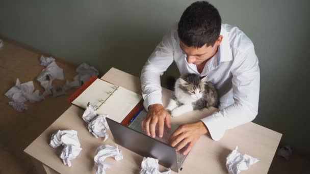 Man Remotely Works at Laptop at Home with a Cat in his Arms (en inglés). Antecedentes del lío — Vídeos de Stock