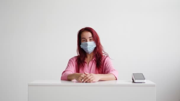 Vrouwelijke beheerder met medisch masker staat achter de receptie en nodigt klanten uit — Stockvideo