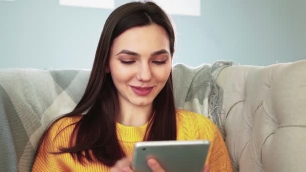 Жінка сидить на затишному дивані посміхається і навчається додаток на цифровому комп'ютері планшеті — стокове відео