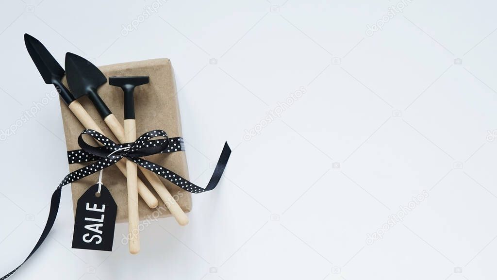 Online shopping for Black Friday.Brown gift box,garden tool with black bow in white peas and black tag on white background, top view, flat lay, copy space. Garden tool sale concept