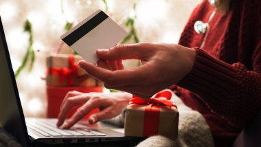 Elinde kredi kartı olan genç bir kadın dizüstü bilgisayarla Noel dekoru ve hediyelerinin arka planına karşı alışveriş yapıyor. Yakın plan. Noel ve Yeni Yıl çevrimiçi alışveriş, kredi kartı ödemeleri.
