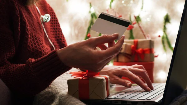 Young woman holding a credit card and makes purchases on a laptop against the background of Christmas decor and gifts, close-up. Christmas and New Years online shopping, credit card payments.