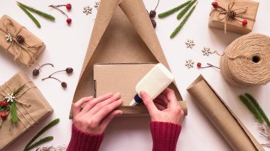 2.Adım adım çevre dostu ve sıfır atık Noel hediyesi paketi için talimat. Kadın hediyeyi kahverengi kağıda sarar ve kenarları yapıştırıcı, üst görünüm, düz uzanma ile mühürler
