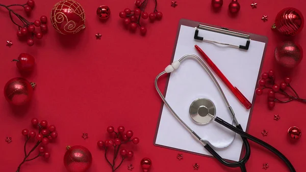 Christmas medical banner, red balls, berries, stars, stethoscope and tablet on red background top view, flat lay.Copyspace. Medicine new year flatly. Doctor writes down the text