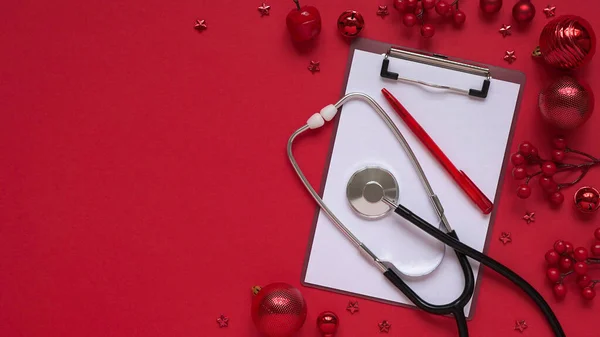 Christmas medical banner, red balls, berries, stars, stethoscope and tablet on red background top view, flat lay.Copyspace. Medicine new year flatly. Doctor writes down the text