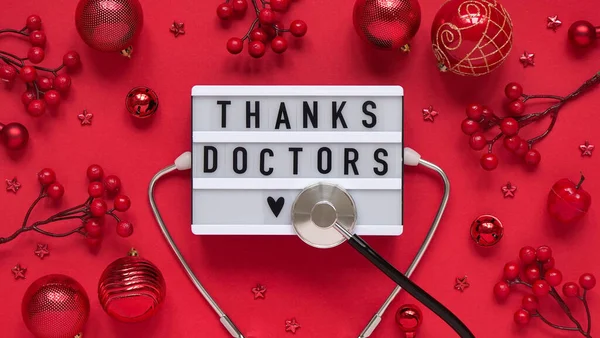 Christmas medical banner,letter board with text thanks doctors,red balls,berries,stars and stethoscope on red background top view,flat lay.Copyspace.Medicine new year flatly.Nameplate lettering