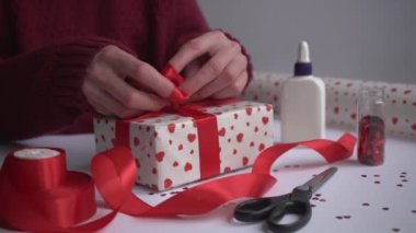 Sevgililer günü hediye paketleme konsepti. Burgundy kazaklı kadın hediyesini beyaz kağıda sarıp bağlıyor. Kırmızı kurdele, kırmızı kurdele, bir rulo ambalaj kağıdı, makas, şeker ve yapıştırıcı..