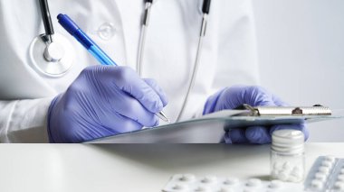 Tablet üzerine reçete yazan doktor ellerinin yakın plan çekimi. Beyaz önlüklü ve mavi eldivenli doktor elinde haplar tutuyor ve mavi kalemle reçete yazıyor. Tıbbi pankart.