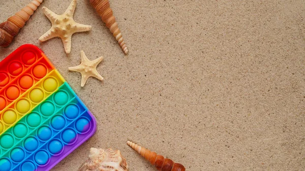 Beach summer vacation banner concept with trendy bright rainbow silicone stress relief toy.Colorful pop it toy,seashells and starfishes lie sand,top view,flatlay,copy space.Fashionable fidgeting game