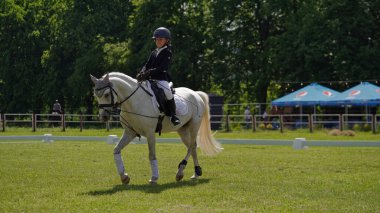 Binicilik spor yarışmasında beyaz atlı kız. Binicilik sınavı. Binicilik yarışmaları, at yarışları. Güneşli yaz gününde ata binen kız jokey. Ukrayna, Kiev 06.04.2021.
