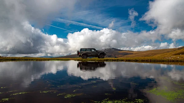 Offroad araba silueti bir dağ sırasındaki küçük bir gölün yakınındaki toprak yolda park edilmiş. Sonbahar renklerini ısıt