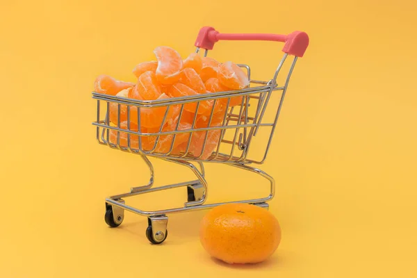 Fresco, suculento, descascado tangerina cunhas em um carrinho de compras e tangerinas ao lado dele. Conceito - na loja online você pode comprar absolutamente qualquer produto com entrega em domicílio. Fundo amarelo. — Fotografia de Stock