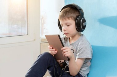 Bir anaokulu çocuğu elinde tablet ve kulaklıkla pencerenin kenarında oturur, müzik dinler, film izler ya da oyun oynar. Yatay fotoğraf