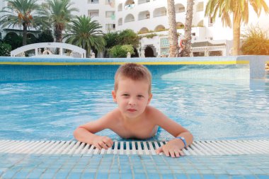 Havuzdaki küçük bir çocuğun portresi şık beyaz bir otelin ve palmiye ağaçlarının arka planına karşı. Çocuk kameraya bakıyor. yatay fotoğraf. Aile tatili